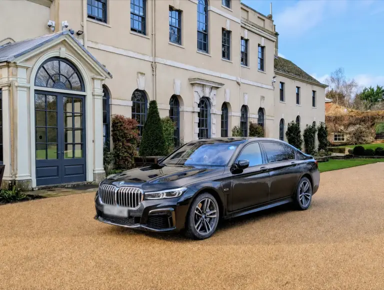 A sleek, black BMW 7 Series M Sport sedan is parked on a gravel driveway in front of a grand, multi-story estate building.