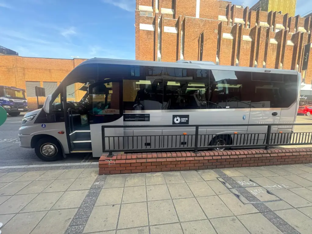 An executive silver midi-coach minibus is parked on a city street next to a brick-based black metal railing, with a prominent square black sticker displaying a white logo and the words "BANKING CIRCLE" on its side panel.